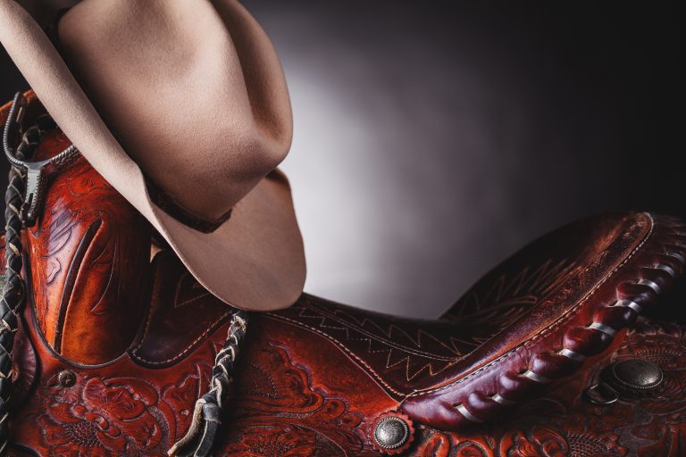 A cowboy hat resting on a deep red-brown saddle in a small studio setting.